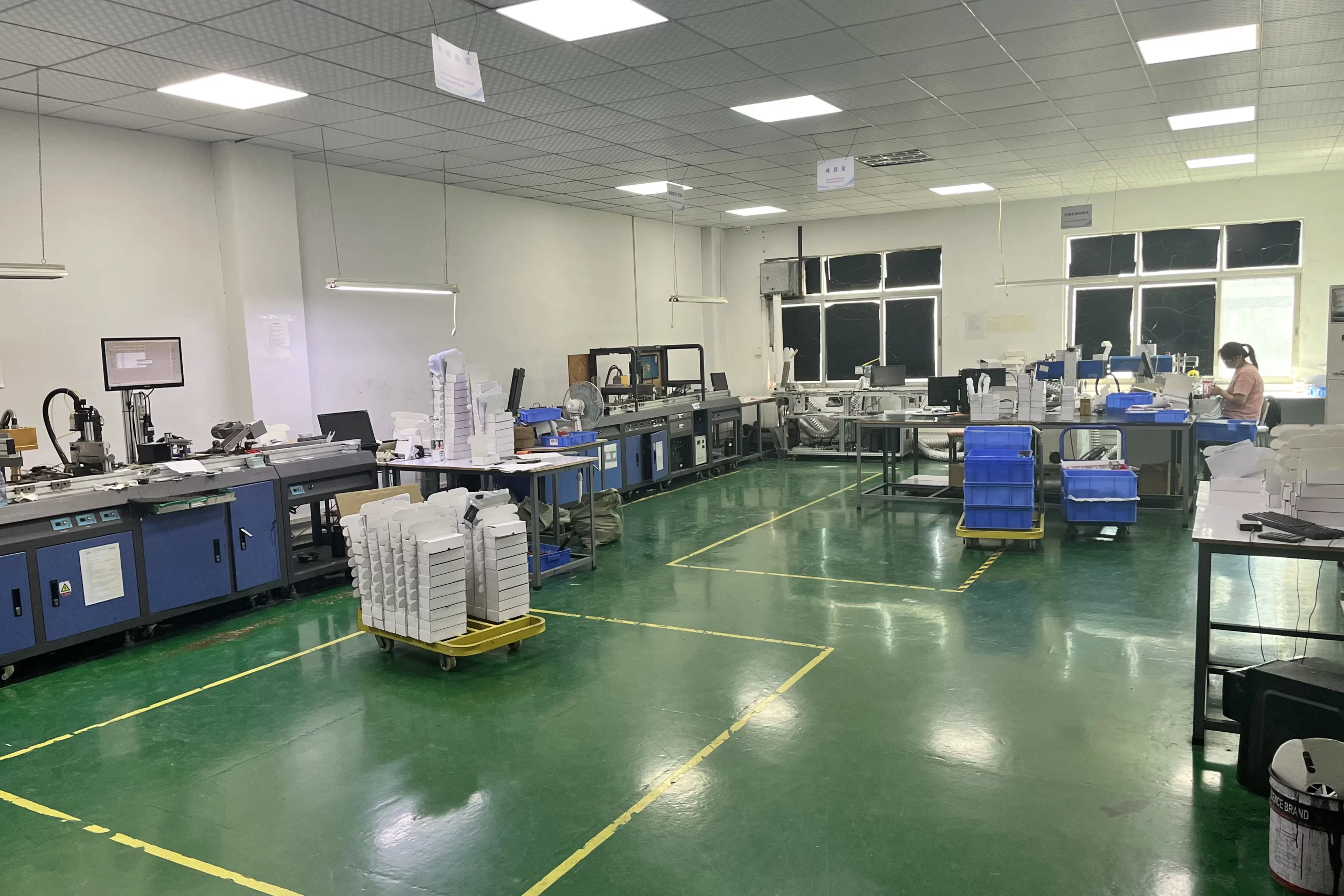 Un pulito, Impianto di produzione organizzato con postazioni di lavoro, attrezzatura, Vassoi impilati, e qualche bidone blu. Una persona sta lavorando in una stazione sul lato destro dell'immagine.
