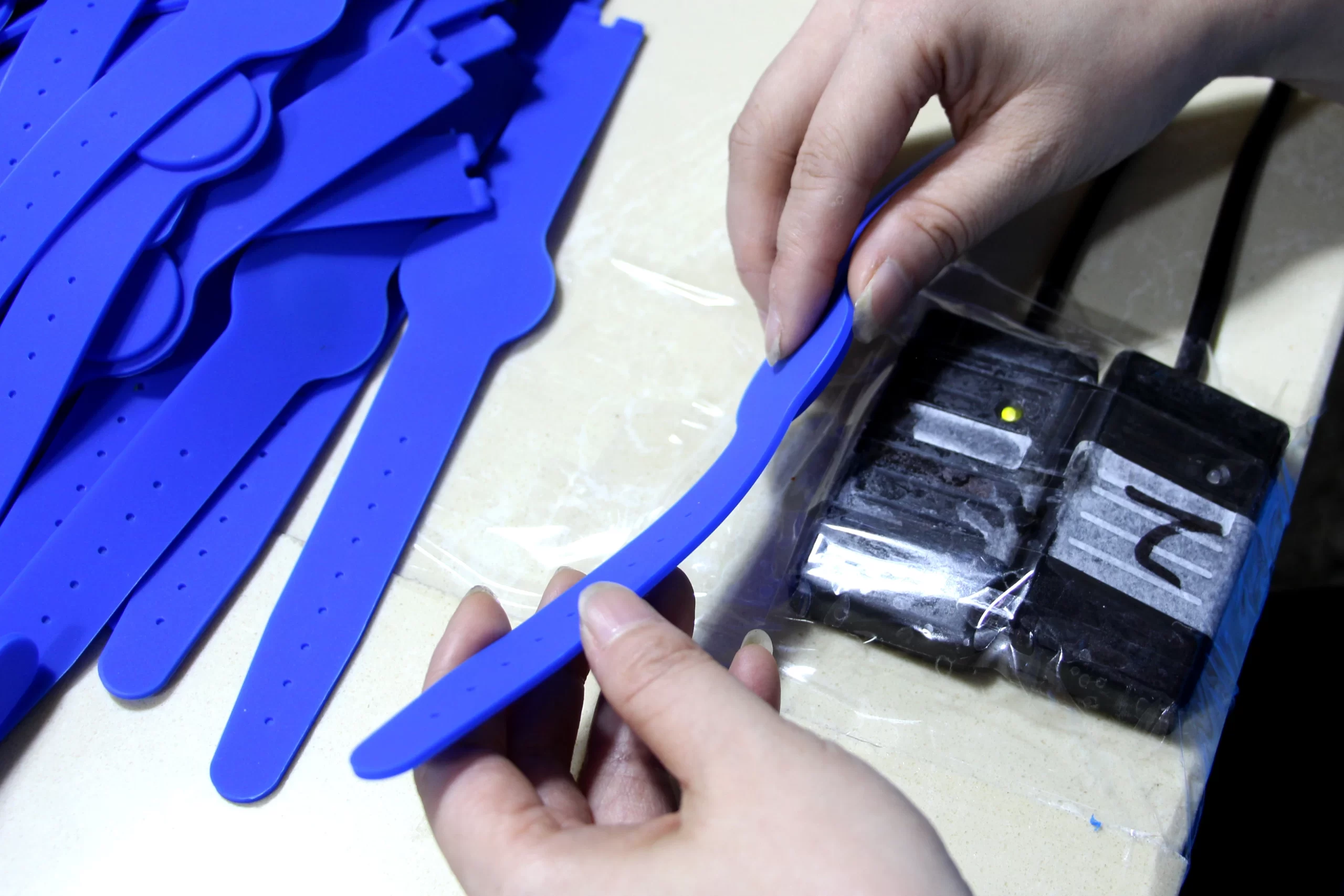 Mani che tengono e ispezionano un braccialetto blu accanto a una pila di braccialetti simili e un dispositivo elettronico avvolto nella plastica.