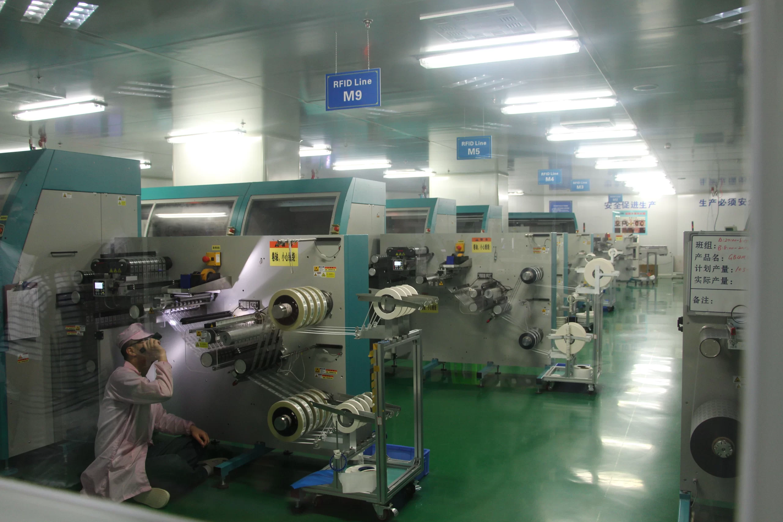 Uomo che lavora in un impianto di produzione high-tech con macchinari e sistemi di trasporto avanzati. La segnaletica per le linee RFID M9 e M5 è visibile. Il lavoratore indossa un camice da laboratorio e un berretto.