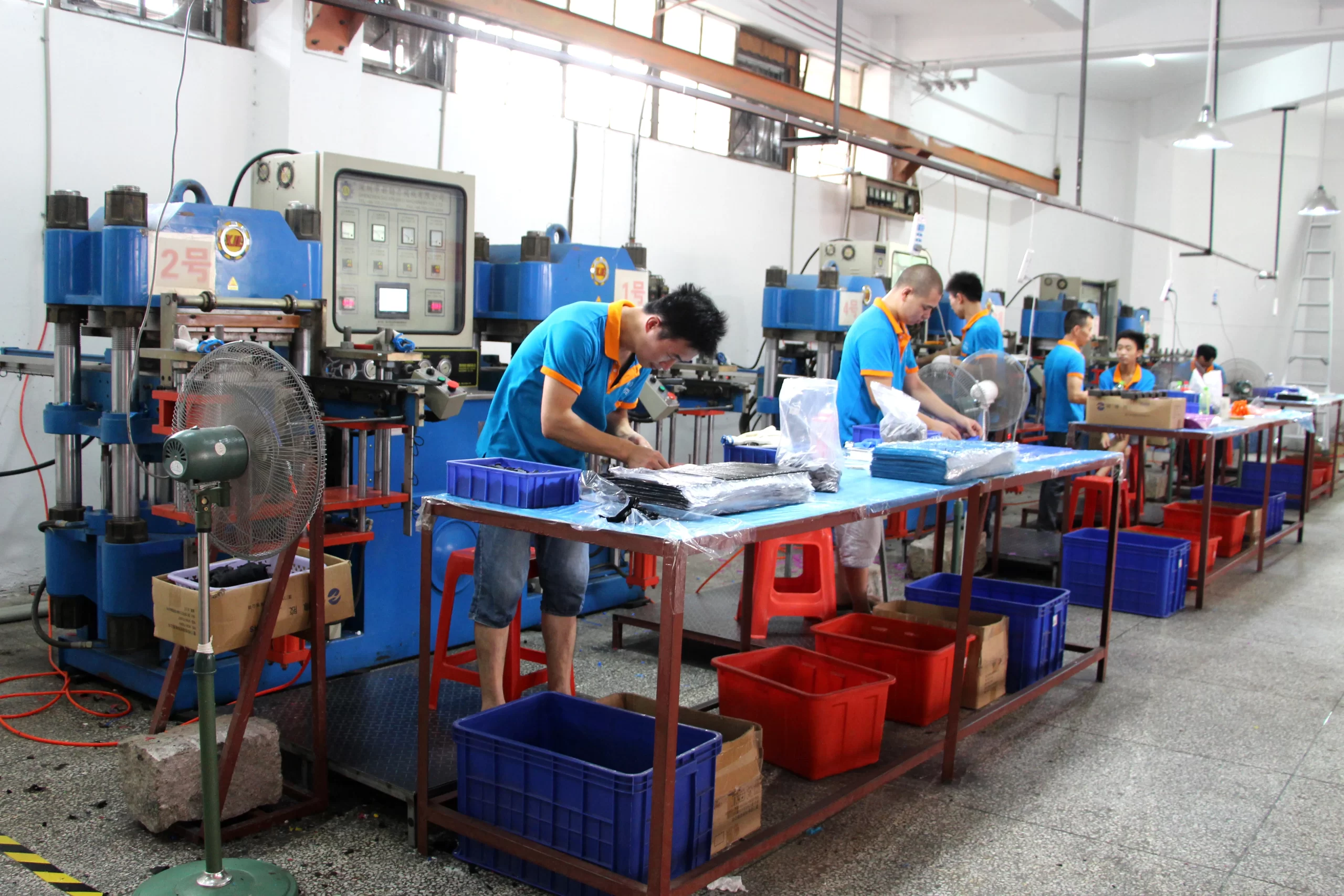 Lavoratori in camicia blu azionano macchinari e maneggiano materiali in un impianto di produzione. Sono visibili vari contenitori di plastica e postazioni di lavoro organizzate.